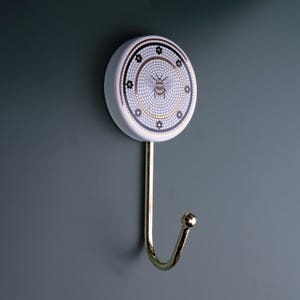 Könnte beinhalten: Weißer Keramikhaken mit einem goldenen Haken und einem Bienendesign. Die Biene ist von einem Mosaikmuster mit schwarzen und goldenen Akzenten umgeben.