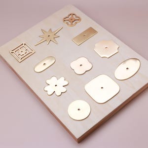May include: A collection of eleven gold-toned metal hardware pieces in various geometric and organic shapes, including stars, circles, squares, and floral patterns, each featuring a single central hole, arranged on a light wooden board.