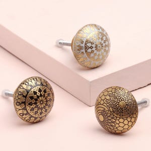 Op de afbeelding: Drie decoratieve kastknoppen met ingewikkelde gouden en zwarte patronen. Eén knop is wit met gouden details. De knoppen hebben zilveren hardware en staan tegen een lichtroze achtergrond. Ideaal om een vleugje elegantie aan meubels toe te voegen.