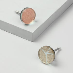 Può includere: Due pomelli decorativi per armadi. Uno è oro rosa con un design geometrico, l'altro è dorato con un design bianco. Entrambi hanno una faccia di vetro circolare trasparente e una vite argentata.