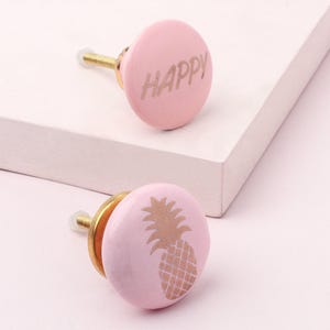 May include: Two pink ceramic cabinet knobs. One knob has the word "HAPPY" in gold lettering. The other knob has a gold pineapple design. Both knobs have gold hardware and are on a white surface.
