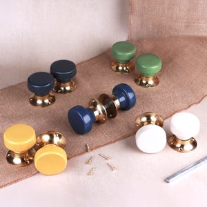 May include: A collection of round door knobs with glossy ceramic tops in yellow, dark blue, green, and white, each featuring a polished brass base with screw holes. A central blue door knob set includes a metal spindle and small screws.
