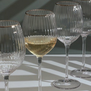 May include: Four clear wine glasses with a ribbed design and gold rims. One glass is partially filled with white wine. The glasses are arranged on a white surface with a soft green background.