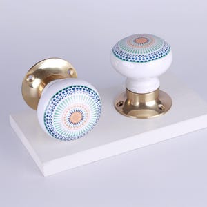 May include: Two white ceramic door knobs with a decorative pattern in blue, green, and peach. The knobs have brass bases and are set against a white background. The design is a circular pattern.