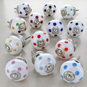 Könnte beinhalten: Eine Sammlung weißer Keramik-Schrankknöpfe mit bunten Polka-Dots. Jeder Knopf hat verschiedenfarbige Punkte, darunter rot, blau, grün, gelb und rosa. Die Knöpfe haben silberfarbene Metallbeschläge und sind auf einer hellen Oberfläche angeordnet.