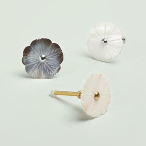 Op de afbeelding: Drie bloemvormige kastknoppen. Eén is donkergrijs en de andere twee zijn wit. Elke knop heeft een zilveren of gouden midden en een schroefdraad voor installatie. De knoppen zijn gemaakt van een parelmoerachtig materiaal.