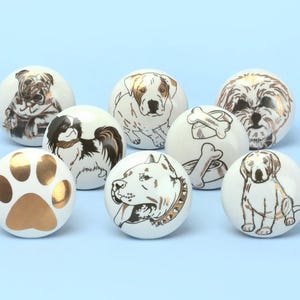 May include: Eight white ceramic cabinet knobs, each decorated with a different dog design in gold and black. The knobs feature various dog breeds, a paw print, and a bone. The knobs are set against a light blue background.