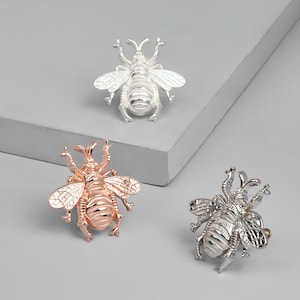 Könnte beinhalten: Drei Bienenbroschen in Silber, Roségold und Gold. Jede Brosche zeigt ein detailliertes Bienendesign mit filigranen Flügeln und einem strukturierten Körper.