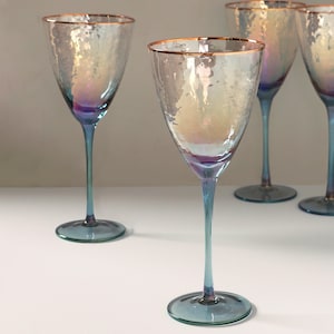 May include: A set of iridescent wine glasses with a textured finish. Each glass has a blue stem and base, and a gold rim. The glasses are arranged on a white surface.
