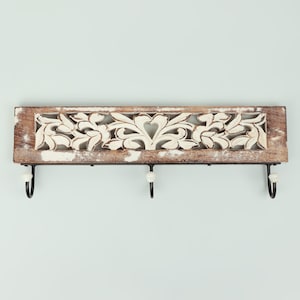 Peut inclure: Un porte-clés mural en bois rustique avec une finition vieillie. La planche rectangulaire présente un motif floral sculpté en blanc et trois crochets en métal noir avec des embouts en céramique blanche. Conçu pour être fixé au mur.