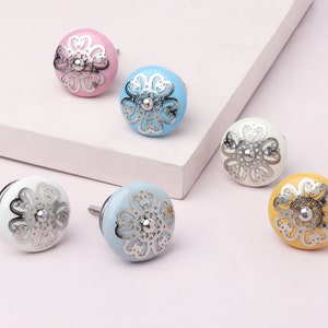 Puede incluir: Seis pomos de armario de cerámica con un diseño floral. Los pomos son de varios colores, incluyendo rosa, azul, blanco y amarillo. Cada pomo tiene un acento de metal plateado en el centro.