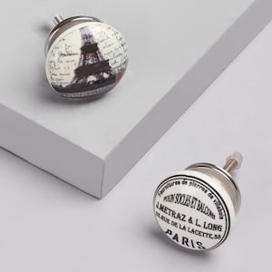 Könnte beinhalten: Zwei dekorative Schrankknöpfe. Ein Knopf zeigt ein Schwarz-Weiß-Bild des Eiffelturms mit Schrift. Der andere Knopf trägt den Text "Fournitures de pierres de villebois POUN SOCLES ET BALCONS J.METRAZ & L. LONG 38 RUE DE LA LACETTE PARIS."
