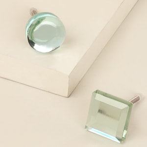 Op de afbeelding: Twee decoratieve glazen kastknoppen. Eén is een heldere, ronde knop en de andere is een vierkante, facetgeslepen knop. Beide knoppen hebben een lichtgroene tint en een zilveren schroef. De knoppen liggen op een lichtbeige oppervlak.