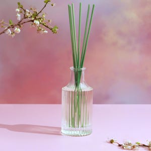 Puede incluir: Un frasco difusor de vidrio transparente con cañas verdes, sobre un fondo degradado rosa y morado. La botella tiene estrías verticales y está parcialmente llena de líquido. Una rama floreciente está en el fondo.