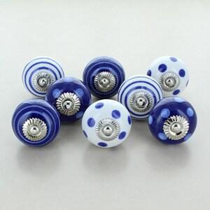 Op de afbeelding: Een verzameling van acht keramische kastknoppen. De knoppen hebben verschillende ontwerpen, waaronder blauw-witte strepen, effen blauw en wit met blauwe stippen. Elke knop heeft een zilverkleurige metalen basis.