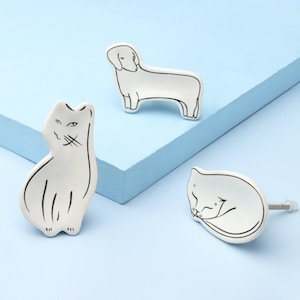 May include: Three white ceramic cabinet knobs with black line drawings of a cat, a sleeping cat, and a dachshund dog. The knobs are on a light blue surface.