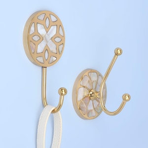 May include: Two decorative wall hooks with a gold finish and inlaid mother-of-pearl accents. One hook holds a white leather strap. The hooks have a circular design with a decorative pattern and are mounted on a light blue wall.