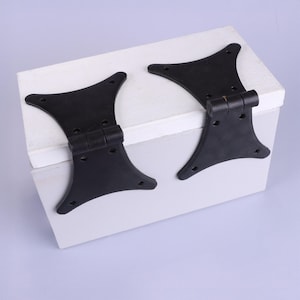 May include: Two black decorative hinges are attached to a white and grey painted wooden box. The hinges have a unique shape with multiple screw holes for secure mounting. The box is on a light purple background.