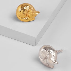 Könnte beinhalten: Zwei katzenförmige Schubladenknöpfe, einer in Gold und einer in Silber. Die Katze ist in einer schlafenden Position zusammengerollt.