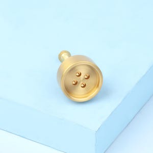 May include: A brass-colored cabinet knob with a cylindrical body and a small, round handle. The knob features four small, crescent-shaped holes on its face. The hardware is set against a light blue background.