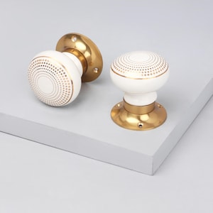 May include: Two white and gold door knobs. The knobs have a round shape with a gold dotted pattern and gold trim. The base and mounting plates are gold. The knobs are on a light gray surface.