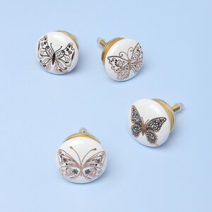 May include: Four white ceramic cabinet knobs with butterfly designs in brown and gold. Each knob has a gold-colored base and a screw for installation. The butterfly patterns vary slightly across the knobs.