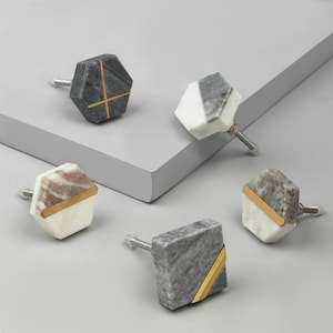 Op de afbeelding: Een verzameling van vijf decoratieve kastknoppen. De knoppen zijn gemaakt van marmer in verschillende kleuren, waaronder grijs, wit en bruin. Sommige knoppen hebben gouden accenten. De knoppen hebben een geometrisch ontwerp.