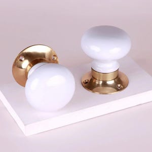 Puede incluir: Dos pomos de puerta con tiradores esféricos de color blanco brillante y liso, montados sobre placas circulares de latón pulido. Cada placa incluye dos orificios visibles para tornillos destinados a su instalación.