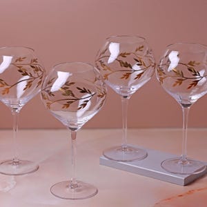 Op de afbeelding: Vier heldere wijnglazen met lange stelen. Elk glas heeft een gouden bladeren ontwerp. De glazen zijn gerangschikt op een lichtgrijs rechthoekig dienblad, tegen een zachtroze achtergrond.
