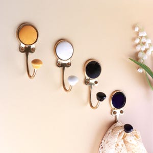 May include: Four wall-mounted hooks with gold-toned metal bases and circular decorative inserts in yellow, white, black, and dark blue. Each hook features a matching colored knob at the end of the curved arm and two small amber-colored accents.