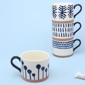 Puede incluir: Tazas de cerámica blanca con asas negras y diseños azules. Una taza presenta un diseño floral, mientras que tres están apiladas, cada una con un patrón geométrico diferente. Las tazas tienen una base marrón, sobre un fondo azul claro.