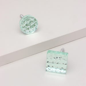 Op de afbeelding: Twee decoratieve kastknoppen. Eén is rond, de andere is vierkant. Beide zijn lichtgroen met een facetontwerp. De knoppen hebben zilverkleurige metalen schroeven. De achtergrond is wit met een lichtgrijze horizontale accent.