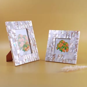 Op de afbeelding: Twee vierkante fotolijsten met een mozaïkpatroon van parelmoer. Elke lijst bevat een foto van een kleurrijk bloemstuk. De lijsten worden ondersteund door houten standaards, tegen een gouden achtergrond.