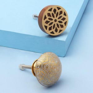 Peut inclure: Deux boutons de meuble décoratifs. L'un est rond avec une base en bois et un motif floral doré. L'autre est un bouton doré et blanc avec un motif orné. Les deux ont des vis argentées et sont posés sur un fond bleu clair.