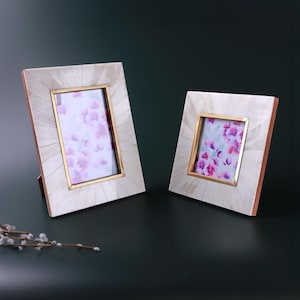 May include: Two decorative photo frames. The frames have a white, textured surface with a gold-colored inner frame. The frames are rectangular and contain a floral print. The frames are set against a dark green background.