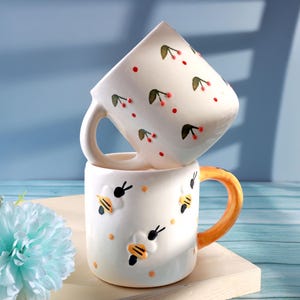 Puede incluir: Dos tazas de cerámica blancas apiladas una encima de la otra. La taza superior tiene un asa y está decorada con puntos rojos y diseños de cerezas verdes y rojas. La taza inferior tiene un asa naranja y diseños de abejas.