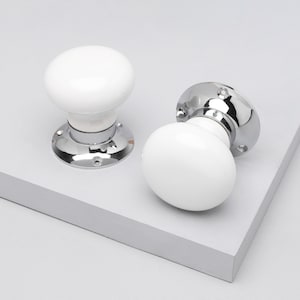 May include: Two white ceramic door knobs with a polished chrome base. The knobs have a rounded shape and are set against a light gray background. The hardware is designed for interior doors.