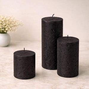 May include: Three black pillar candles of different heights. The candles have a textured surface and a wick. A small white vase with white flowers is in the background.