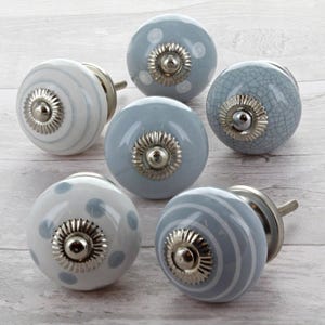 Op de afbeelding: Zes decoratieve keramische kastknoppen in blauw en wit. Ontwerpen omvatten stippen, strepen en een craquelé-patroon. Elke knop heeft een zilverkleurige metalen basis en schroef.