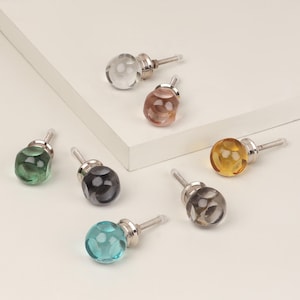 Può includere: Una collezione di sette pomelli per armadi in vetro di vari colori, tra cui trasparente, ambra, verde e blu. Ogni pomello ha un design rotondo e sfaccettato e una base in metallo argentato. I pomelli sono disposti su una superficie bianca.