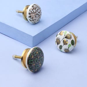 May include: Three decorative ceramic cabinet knobs with brass fittings. The knobs display distinct patterns: a gold and black tree design, a green and black geometric pattern, and a floral design with gold and green accents. Set against a light blue backdrop.