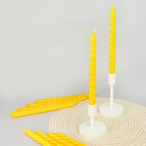 Könnte beinhalten: Gelbe gedrehte Kerzen, einige in weißen Kerzenhaltern, vor neutralem Hintergrund. Mehrere Kerzen sind zu sehen, einige stehen aufrecht, andere liegen. Die Kerzen haben ein gedrehtes Design.
