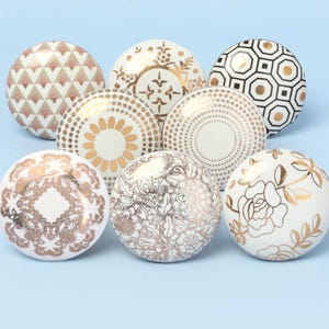 May include: Eight decorative cabinet knobs with diverse patterns. Designs include geometric shapes, floral motifs, and dotted patterns in gold and white. The knobs are arranged on a light blue surface.