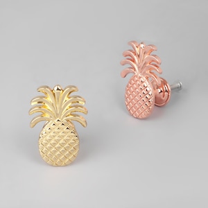 May include: Two pineapple-shaped cabinet knobs. One is gold-colored, and the other is rose gold. Both knobs have a textured surface and a detailed crown. The knobs are designed for furniture and home decor.