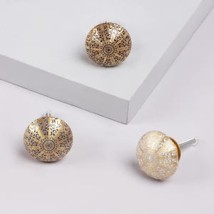 Può includere: Tre pomelli decorativi per mobili con finitura oro e bronzo. Ogni pomello presenta un intricato design floreale circolare. Un pomello è su una vite argentata. I pomelli sono esposti su una superficie bianca.
