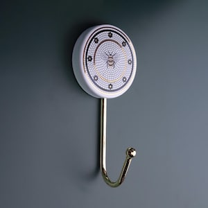 May include: A decorative wall hook with a white, round ceramic face featuring a mosaic design with a bee in the center. The hook has a gold-colored metal stem and a curved hook end. The design includes black and gold accents.