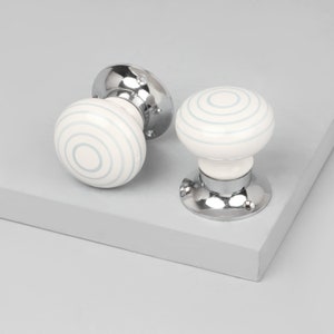 May include: Two round cabinet knobs featuring a white ceramic surface with concentric light blue circular lines. Each knob is attached to a polished chrome metal base with a circular mounting plate that includes two small screw holes for installation.