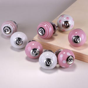 Puede incluir: Una colección de tiradores decorativos para armarios en varios diseños. Los tiradores presentan una mezcla de colores blanco y rosa con patrones como rayas, lunares y efectos craquelados. Cada tirador tiene una base de metal plateado.