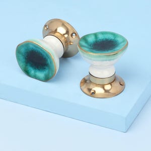 May include: Two decorative door knobs with a teal and white ceramic design. The knobs have a gold trim and base, set against a light blue background. The ceramic portion has a unique, organic shape.