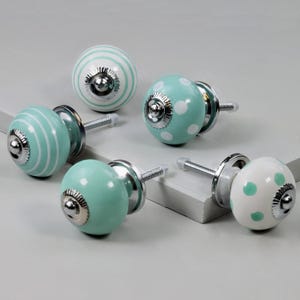 May include: A collection of decorative ceramic cabinet knobs in various designs. The knobs feature a mix of colors, including white, mint green, and silver. Some knobs have stripes, while others have polka dots. Each knob has a metal base and screw.